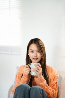 Oturma odasında, pencerenin yanında bir fincan sıcak içecekle rahatlayan Asyalı kadın. Rüya gören bayan, stres olmadan sakin havanın tadını çıkar, yalnız olmanın tadını çıkar. Evde rahat bir sonbahar..