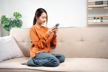 Gülümseyen Asyalı kadın turuncu kazaklı akıllı telefon kullanıyor evde çalışıyor, Asyalı güzel kız hygge oturma odasında dinleniyor. Teknoloji insanları dijital çevrimiçi sosyal medya pazarlama afişini bağlıyor