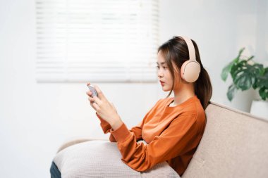 Genç Asyalı oyuncu kadın kanepede uzanıyor, kulaklık takıyor ve elinde akıllı telefon var. Oyun deneyiminin tadını çıkarıyor. Zafer havası için yarışma. İnsanlar evlerinde boş zaman geçiriyorlar..