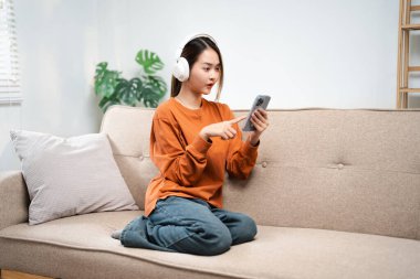 Mutlu Asyalı kadın cep telefonundan müzik dinliyor evdeki bir kanepede oturuyor. Sabahları kulaklıkla rahatlayan gülümseyen kız, dinlenme zamanı. Boşluğu kopyala.