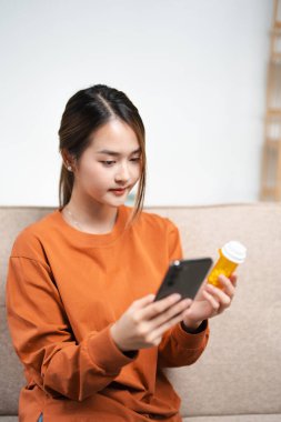 Sağlık hizmetlerinde çalışan Asyalı genç bir kadın okumak için akıllı telefon kullanıyor, ilaç reçetelerini araştırıyor, internetteki bilgiler hakkında ilaç etiketi metni yazıyor, talimatlar yan etkiler, tıbbi konsept.