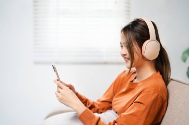 Mutlu Asyalı kadın cep telefonundan müzik dinliyor evdeki bir kanepede oturuyor. Sabahları kulaklıkla rahatlayan gülümseyen kız, dinlenme zamanı. Boşluğu kopyala.
