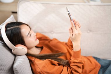 Mutlu Asyalı kadın cep telefonundan müzik dinliyor evdeki bir kanepede oturuyor. Sabahları kulaklıkla rahatlayan gülümseyen kız, dinlenme zamanı. Boşluğu kopyala.