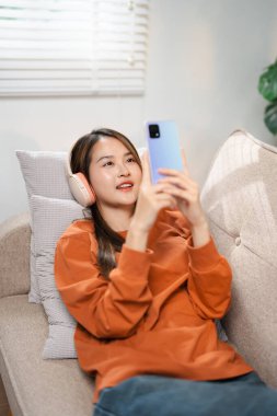 Asyalı kadın telefon için ses çalar uygulaması kullanıyor. Cep telefonundan müzik dinleyen mutlu bir kadın evdeki rahat koltukta oturuyor. Sabahları kulaklıkla rahatlayan gülümseyen kız, dinlenme zamanı. Boşluğu kopyala.