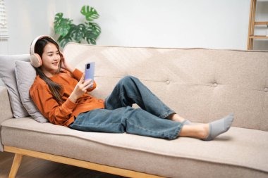 Asyalı kadın telefon için ses çalar uygulaması kullanıyor. Cep telefonundan müzik dinleyen mutlu bir kadın evdeki rahat koltukta oturuyor. Sabahları kulaklıkla rahatlayan gülümseyen kız, dinlenme zamanı. Boşluğu kopyala.