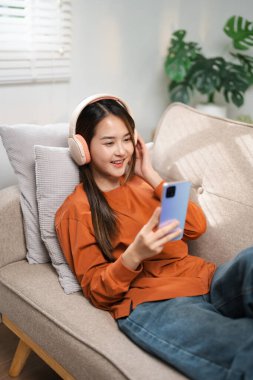 Mutlu kadın, kulaklık ve akıllı telefon oturma odasında sosyal medya uygulaması, sesli kitap ya da kanepede radyo yayını. Genç kız, mobil müzik ve dinlenme odasında ses, yayın ve şarkı dinleme.