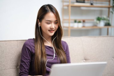 Kulaklıklı genç Asyalı kadın. Yazıyor ve not alıyor. İnternetteki laptop ekran çalışmasına bakın. Kulaklıklı mutlu kadın bilgisayarla uzaktan çalışıyor. Eğitim kavramı.