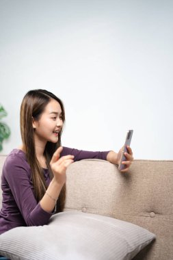 Asyalı kadın, elinde telefonla koltukta oturup cep telefonunun ekranına bakarak akıllı telefon görüntülü sohbet uygulaması kullanarak mobil sohbet uygulamasında uzaktan arama mesafesini gösteriyor. uzak web kamera çağrısı.