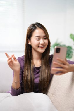 Asyalı kadın, elinde telefonla koltukta oturup cep telefonunun ekranına bakarak akıllı telefon görüntülü sohbet uygulaması kullanarak mobil sohbet uygulamasında uzaktan arama mesafesini gösteriyor. uzak web kamera çağrısı.