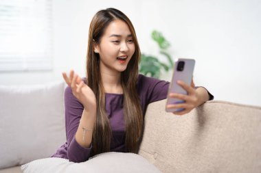 Asyalı kadın, elinde telefonla koltukta oturup cep telefonunun ekranına bakarak akıllı telefon görüntülü sohbet uygulaması kullanarak mobil sohbet uygulamasında uzaktan arama mesafesini gösteriyor. uzak web kamera çağrısı.