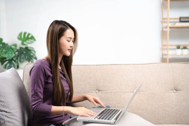 Pozitif Asyalı kız kanepeye oturup, modern dizüstü bilgisayarla sınırsız kablosuz interneti tarıyor, evden gelen bilgisayar daktilo yazıları ve teknoloji konsepti üzerine mutlu genç kadın serbest çalışıyor..