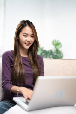 Pozitif Asyalı kız kanepeye oturup, modern dizüstü bilgisayarla sınırsız kablosuz interneti tarıyor, evden gelen bilgisayar daktilo yazıları ve teknoloji konsepti üzerine mutlu genç kadın serbest çalışıyor..