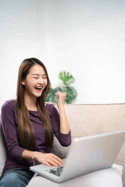 Heyecanlı Asyalı kadın online galibiyeti kutluyor. Evde dizüstü bilgisayara bakıyor. Kazanan el kaldırıyor. Başarıyı kutlayan yeni bir iş fırsatı elde ediyor. Bilgisayarda maç veya spor izliyor..