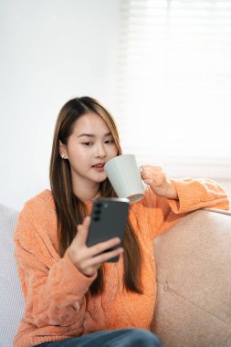 Genç mutlu kadın cep telefonu kullanıyor kahve içiyor evde dinleniyor. Gülümseyen güzel bayan elinde akıllı telefonla gezintiye çıkıyor, sosyal medya mesajlarını kontrol ediyor, oyun oynuyor ya da internetten alışveriş yapıyor..