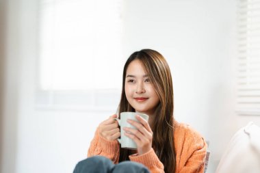 Oturma odasında, pencerenin yanında bir fincan sıcak içecekle rahatlayan Asyalı kadın. Rüya gören bayan, stres olmadan sakin havanın tadını çıkar, yalnız olmanın tadını çıkar. Evde rahat bir sonbahar..