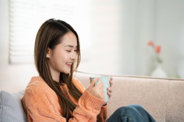 Mutlu Asyalı kadın tatilde sıcak kahve ya da çay içerek evdeki koltukta rahatlıyor, rahat sahne, gülen güzel kadın sonbahar kışında sıcak çay içiyor. Boşluğu kopyala.