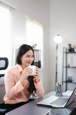 Asyalı serbest çalışan rahat bir kadın laptopun önünde kahve içip rahatlıyor, çalışıyor ya da evden uzakta ders çalışıyor, fincandan içiyor ve dinleniyor..
