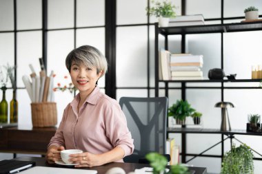 Ofiste elinde kahve fincanıyla gülümseyen başarılı bir iş kadını. Kendine güvenen Asyalı iş kadını ofiste oturup kahve içiyor..