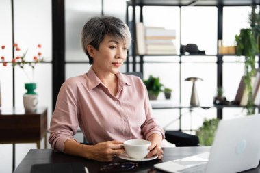 Ofiste elinde kahve fincanıyla gülümseyen başarılı bir iş kadını. Kendine güvenen Asyalı iş kadını ofiste oturup kahve içiyor..