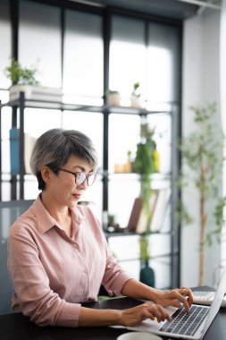 Ev ofisinde çalışan gözlüklü orta yaşlı bir iş kadını kopya alanı olan bir bilgisayar kullanıyor. Ofiste dizüstü bilgisayarda çalışan Asyalı kadın..