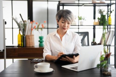 Beyaz gömlek giyen, tablet kullanan, rahat çalışma alanında oturan olgun Asyalı iş kadını. Ofiste duran kendine güvenen bir iş adamı..