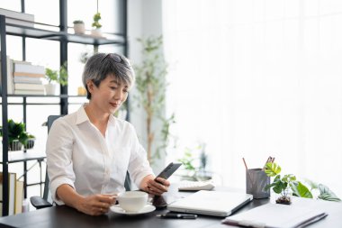 Evde dizüstü bilgisayarla çalışırken cep telefonu kullanan son sınıf öğrencisi mutlu bir kadın. Asyalı havalı yaşlı kadın akıllı telefonuyla mesajlaşıyor. Güzel şık, yaşlı kadın cep telefonunda geziniyor..