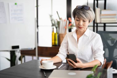 Evde dizüstü bilgisayarla çalışırken cep telefonu kullanan son sınıf öğrencisi mutlu bir kadın. Asyalı havalı yaşlı kadın akıllı telefonuyla mesajlaşıyor. Güzel şık, yaşlı kadın cep telefonunda geziniyor..
