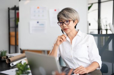 Kendine güvenen yaşlı bir kadın, modern ofiste çalışan dizüstü bilgisayarı kullanan bir yönetici. Şık gözlükler takan iş kadını e-postaları kontrol ediyor. Teknoloji konsepti
