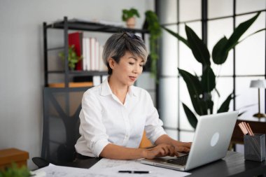 Çekici, kısa saçlı, orta yaşlı, beyaz tişörtlü bir kadın. IT proje yöneticisi iş yerinde oturuyor, yeşil ofisteki dizüstü bilgisayarda çalışıyor.