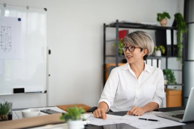 Ofis masasında oturan kadın mimar planlar, kadastro haritaları ve şehir planlarıyla çalışıyor. Asyalı kadın bilgi okuyor ve yeni bir mimari proje geliştiriyor