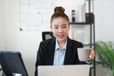 Ev ofisinin masasında elinde laptop ve belgelerle kahve molası veren gülümseyen bir kadın, parlak ve rahat bir ortamda çalışıyor..