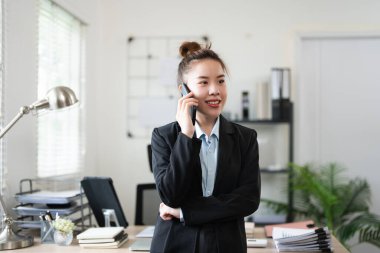 Genç, meşgul bir iş kadını modern ofiste telefonla konuşuyor. Asyalı iş kadını şirket satış müdürü takım elbise giyiyor cep telefonuyla arama yapıyor iş yerinde duruyor.