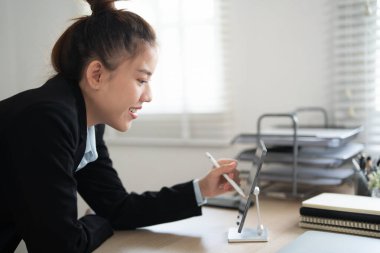 Siyah takım elbiseli, mutlu, mutlu, çalışan genç bir iş kadını açık modern ofisteki dizüstü bilgisayarla çalışma masasında kalem tutuyor. Başarı kariyer konsepti