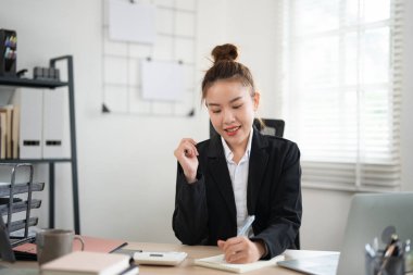 Siyah takım elbiseli, mutlu, mutlu, çalışan genç bir iş kadını açık modern ofisteki dizüstü bilgisayarla çalışma masasında kalem tutuyor. Başarı kariyer konsepti
