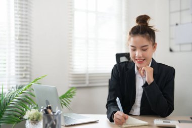 Masasında oturan Asyalı genç bir iş kadını. Kalem tutuyor. Kapalı bir ofiste not defteri yazıyor.