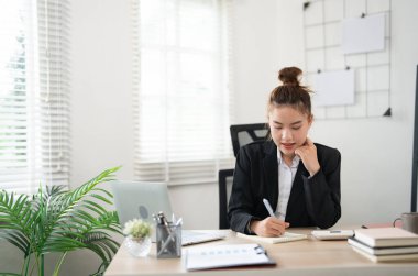 Siyah takım elbiseli, mutlu, mutlu, çalışan genç bir iş kadını açık modern ofisteki dizüstü bilgisayarla çalışma masasında kalem tutuyor. Başarı kariyer konsepti