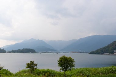 Bunlar Japonya 'da yuva yapmış küçük bir köy olan Fujiyoshida' nın yuvarlanan tepeleri. Bulutlar yanlarından süzülüyor gibi görünüyor. Kawaguchi Gölü aşağıda gösterilir, genellikle sakin görünür..
