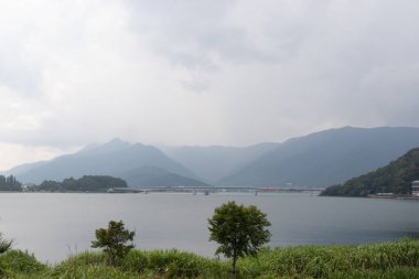 Bunlar Japonya 'da yuva yapmış küçük bir köy olan Fujiyoshida' nın yuvarlanan tepeleri. Bulutlar yanlarından süzülüyor gibi görünüyor. Kawaguchi Gölü aşağıda gösterilir, genellikle sakin görünür..