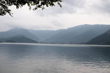 Bunlar Japonya 'da yuva yapmış küçük bir köy olan Fujiyoshida' nın yuvarlanan tepeleri. Bulutlar yanlarından süzülüyor gibi görünüyor. Kawaguchi Gölü aşağıda gösterilir, genellikle sakin görünür..