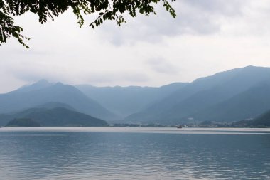 Bunlar Japonya 'da yuva yapmış küçük bir köy olan Fujiyoshida' nın yuvarlanan tepeleri. Bulutlar yanlarından süzülüyor gibi görünüyor. Kawaguchi Gölü aşağıda gösterilir, genellikle sakin görünür..