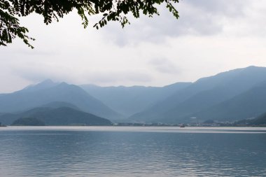 Bunlar Japonya 'da yuva yapmış küçük bir köy olan Fujiyoshida' nın yuvarlanan tepeleri. Bulutlar yanlarından süzülüyor gibi görünüyor. Kawaguchi Gölü aşağıda gösterilir, genellikle sakin görünür..
