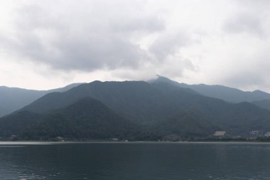 Bunlar Japonya 'da yuva yapmış küçük bir köy olan Fujiyoshida' nın yuvarlanan tepeleri. Bulutlar yanlarından süzülüyor gibi görünüyor. Kawaguchi Gölü aşağıda gösterilir, genellikle sakin görünür..