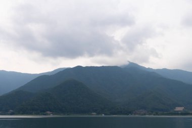 Bunlar Japonya 'da yuva yapmış küçük bir köy olan Fujiyoshida' nın yuvarlanan tepeleri. Bulutlar yanlarından süzülüyor gibi görünüyor. Kawaguchi Gölü aşağıda gösterilir, genellikle sakin görünür..