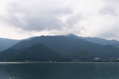 Bunlar Japonya 'da yuva yapmış küçük bir köy olan Fujiyoshida' nın yuvarlanan tepeleri. Bulutlar yanlarından süzülüyor gibi görünüyor. Kawaguchi Gölü aşağıda gösterilir, genellikle sakin görünür..