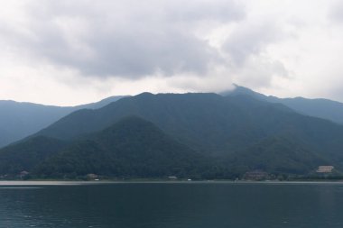 Bunlar Japonya 'da yuva yapmış küçük bir köy olan Fujiyoshida' nın yuvarlanan tepeleri. Bulutlar yanlarından süzülüyor gibi görünüyor. Kawaguchi Gölü aşağıda gösterilir, genellikle sakin görünür..
