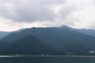 Bunlar Japonya 'da yuva yapmış küçük bir köy olan Fujiyoshida' nın yuvarlanan tepeleri. Bulutlar yanlarından süzülüyor gibi görünüyor. Kawaguchi Gölü aşağıda gösterilir, genellikle sakin görünür..