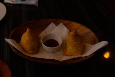 Bu güzel samosalar, aralarında sos bulunan ahşap bir tabağın üzerinde oturuyorlar. Bu kızartılmış kahverengi kızılderili çadırı görünümlü nesneler bir Hint lezzeti. Aperatif olarak sunulan eşyalar beyaz bir peçetenin üzerinde duruyor.. 