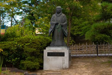 Geleneksel Japon cüppeli bir adamın bronz heykeli taş bir kaidede duruyor. Heykel, huzurlu bir bahçe ortamında yemyeşil ağaçlar ve yapraklarla çevrili. Arka planda alçak, ahşap bir çit görünüyor..