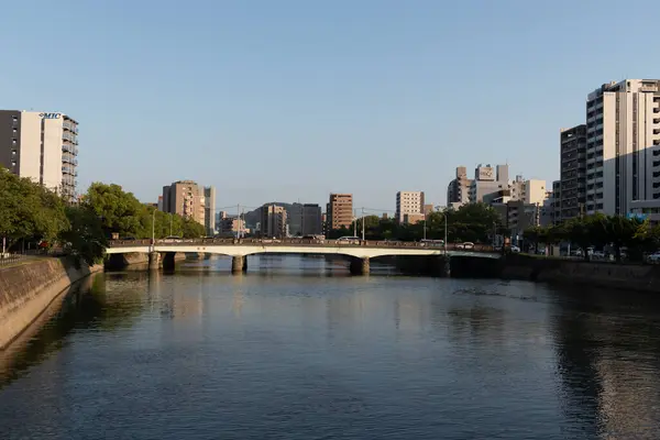 Geniş bir nehir boyunca uzanan canlı bir şehir berrak mavi bir gökyüzünün altında yansıtıcı sularını kaplayan uzun bir köprü. Modern binalar ve yemyeşil ağaçlar nehir kıyılarını kaplayarak kentsel gelişim ve doğal huzurun uyumlu bir karışımını oluşturuyor..
