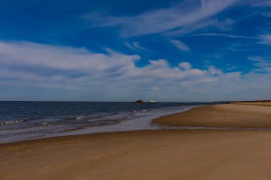Uzak beyaz tekne ve kara gemi kalıntıları parçalı bulutlu bir gökyüzünün altında engin, koyu mavi bir denizde oturuyorlar. Ön planda Sandy Shore var. Anımsatıcı kıyı manzarası.